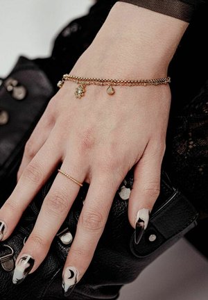 Hand mit schwarz-weißem Marmor-Nageldesign, goldener Kettenring am Finger, Charm-Armband, hält schwarze Lederhandtasche mit silbernen Nieten.