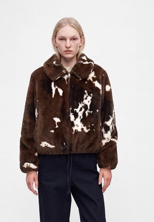 Personne blonde portant une veste en fausse fourrure marron et blanche à motif avec un col et un pantalon foncé sur fond uni.