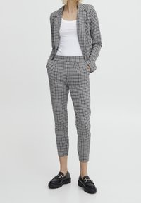 Kobieta w szarym i czarnym w kratę żakiecie oraz dopasowanych spodniach, białym topie na ramiączkach i czarnych loafersach z złotym łańcuszkiem.