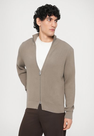 Μπεζ ριμπ ζακέτα με φερμουάρ και ψηλό γιακά, με λείο φερμουάρ και εφαρμοστά μανσέτα, φορεμένη πάνω από λευκό μπλουζάκι και σκούρο παντελόνι.