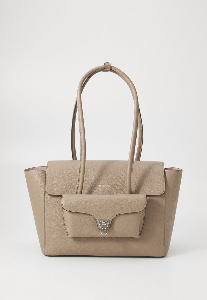 Sac à main en cuir beige avec une forme structurée, dotée de deux poignées, d'une poche avant et d'accents en métal argenté. Texture lisse et design minimaliste.