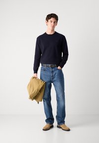 Navy pullover, light blue jeans, tan suede shoes, black belt. Holding a khaki jacket, casual fit, plain design, textured fabric.