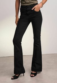 Jeans flares neri con vita alta, caratterizzati da una chiusura a doppio bottone, tasche laterali e una texture liscia. Abbinati a sandali con la punta aperta.