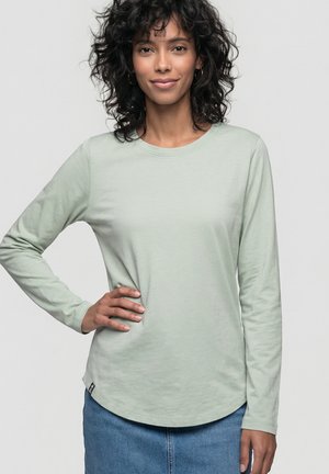 Frau mit lockigem Haar, die ein hellgrünes Langarmshirt und einen blauen Jeansrock trägt, steht mit einer Hand in der Hüfte.