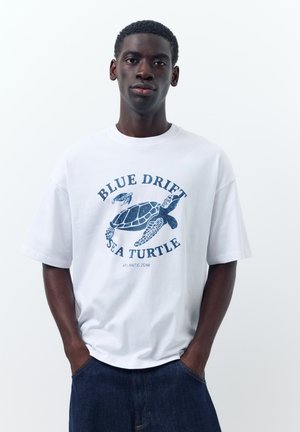 Mladý muž nosí biele tričko s modrým motívom morskej korytnačky a textom „Blue Drift Sea Turtle Atlantic Zone“, ruky v tmavých džínsových vreckách.