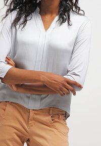 Blusa grigia chiara con scollo a V, maniche a ¾ e vestibilità rilassata, abbinata a pantaloni beige. Tessuto morbido, dettagli minimi e design casual.