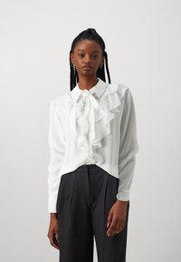 Blusa blanca con fruncido en el frente, cuello con lazo y mangas largas, hecha de un material suave. Combinada con pantalones de corte oscuro en gris.