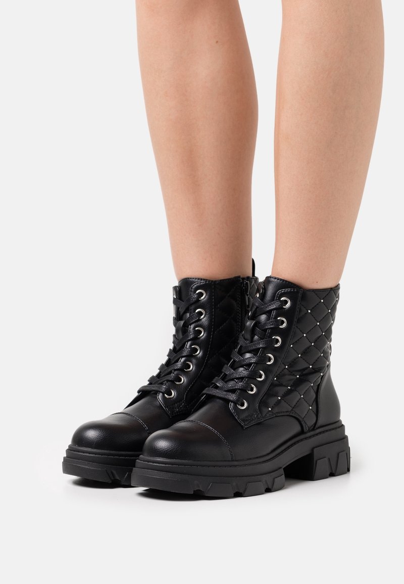 Bottines montantes en cuir noir à lacets avec côtés matelassés et semelles épaisses, portées sur des jambes nues sur fond blanc.
