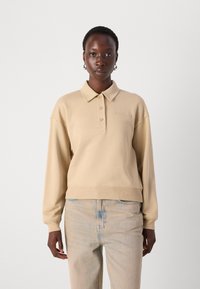 Felpa in cotone beige con colletto, patta a bottoni corta e polsini a coste. Presenta il logo "LACOSTE" in rilievo sul petto. Vestibilità casual.