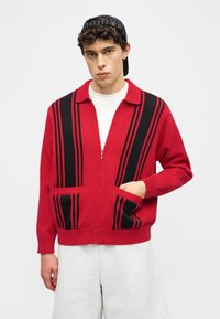 Νεαρός άνδρας που φοράει κόκκινο πουλόβερ με φερμουάρ και μαύρες κατακόρυφες ρίγες, λευκό μπλουζάκι από μέσα, ανοιχτό γκρι σορτς και μαύρο καπέλο με ρίγες φοράει ανάποδα.