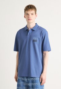 Blå polo med korta ärmar, mjukt tyg, med en mörk fyrkantig logotyp på vänster bröst. Matchas med en rutig blå kjol.