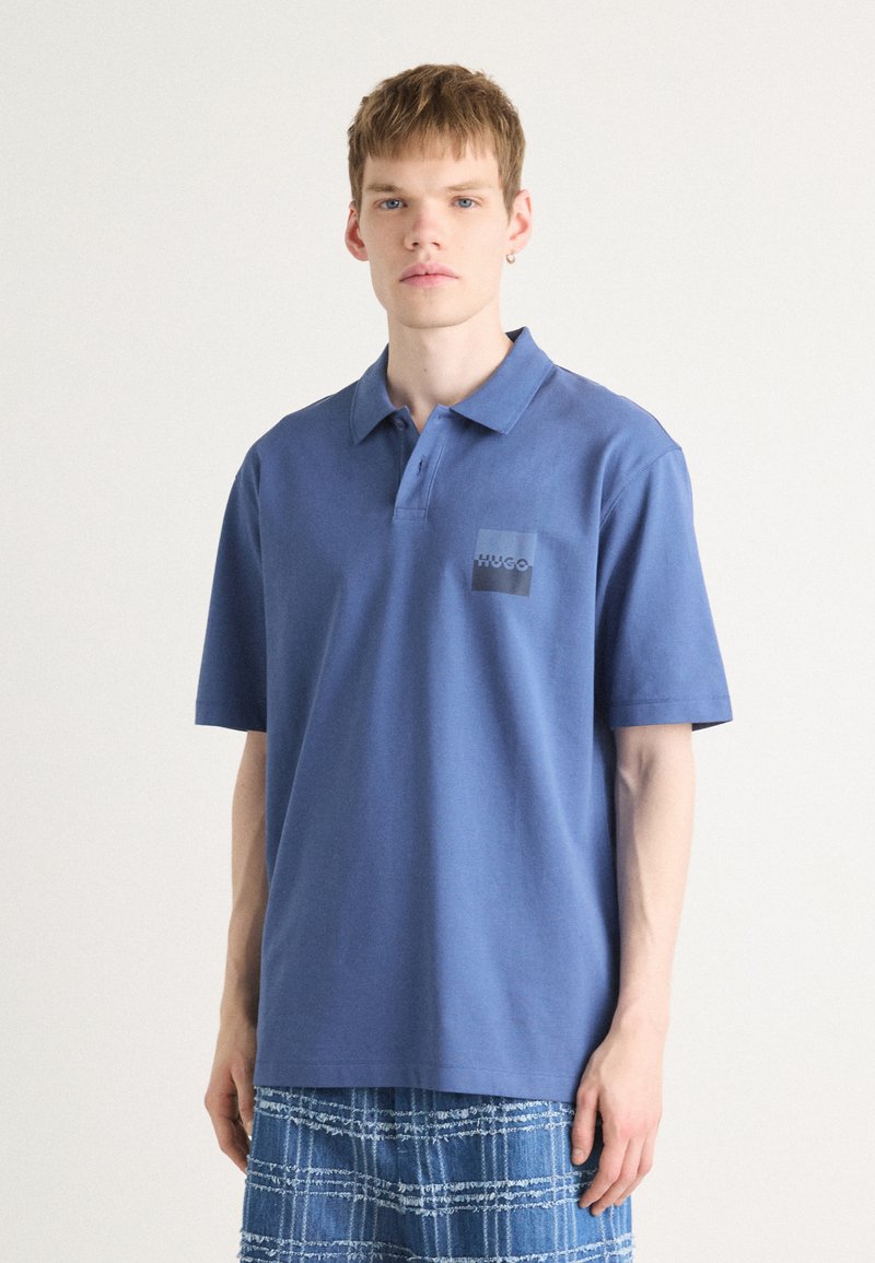 Blå polo med korta ärmar, mjukt tyg, med en mörk fyrkantig logotyp på vänster bröst. Matchas med en rutig blå kjol.