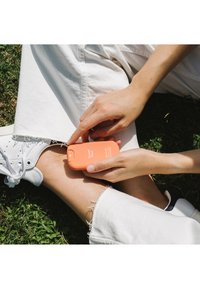 Contenedor cosmético naranja etiquetado "Sunset Fleur" sostenido en una mano, con pantalones blancos y zapatillas visibles, sobre hierba verde.