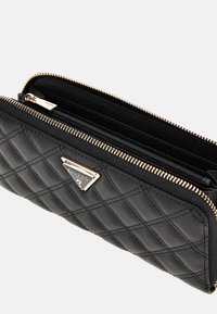 Black quilted leather wallet featuring a gold zip closure and a prominent triangular Guess logo. Interior includes multiple compartments.