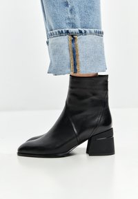 Bottine en cuir noir avec un bout pointu et un talon bloc épais. La bottine a une texture lisse et un design minimaliste.