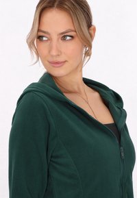 Felpa verde foresta con zip realizzata in morbido materiale felpato, dotata di cappuccio e di un design aderente con sottili dettagli di texture lungo le cuciture.