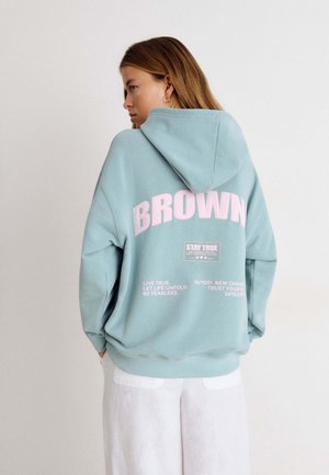 Frau mit langen Haaren, die einen hellblauen Hoodie mit "BROWN" und motivierendem Text auf dem Rücken trägt, kombiniert mit weißen Hosen, von der Seite betrachtet.