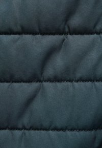 Tissu rembourré en bleu sarcelle avec des lignes de couture diagonales, présentant une texture lisse et un léger éclat. Pas de matériel ou de motifs visibles.