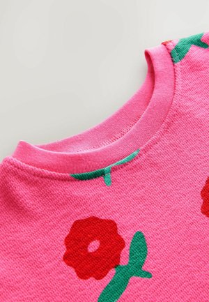 Ružová mikina s textúrovaným povrchom, zdobená veľkými červenými kvetmi a zelenými listami v pruhu okolo výstrihu a ramien.