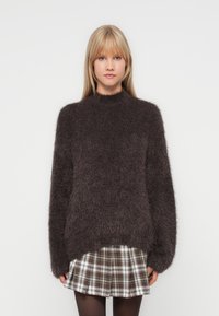 Donkerbruin harige pullover trui met een hoge halslijn en een losse pasvorm, gecombineerd met een geruite rok. De textuur van de stof zorgt voor een gezellige uitstraling.
