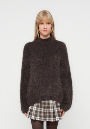 Donkerbruin harige pullover trui met een hoge halslijn en een losse pasvorm, gecombineerd met een geruite rok. De textuur van de stof zorgt voor een gezellige uitstraling.