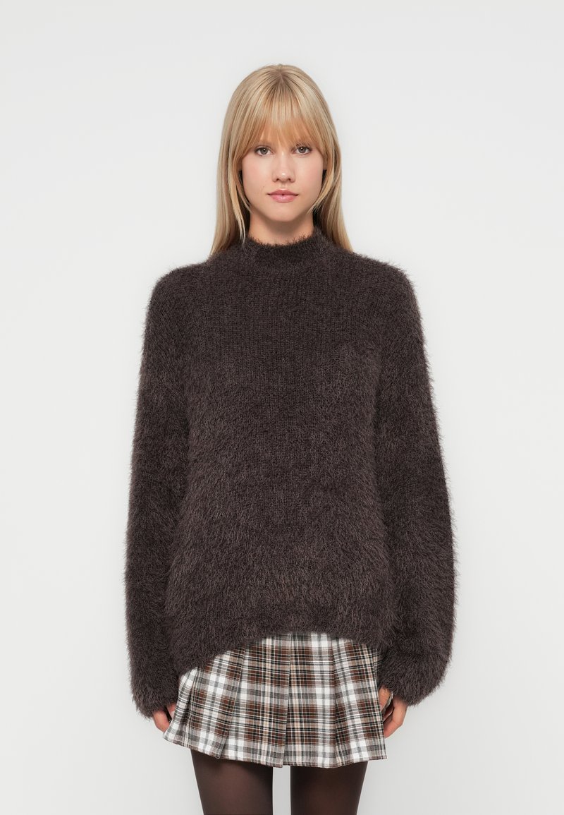 Donkerbruin harige pullover trui met een hoge halslijn en een losse pasvorm, gecombineerd met een geruite rok. De textuur van de stof zorgt voor een gezellige uitstraling.