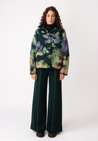 Jakna iz volnenega mešanega materiala v zeleni barvi s cvetličnim vzorcem v vijolični in oranžni barvi, z kapuco in velikimi gumbi, združena s širokimi zelenimi hlačami.