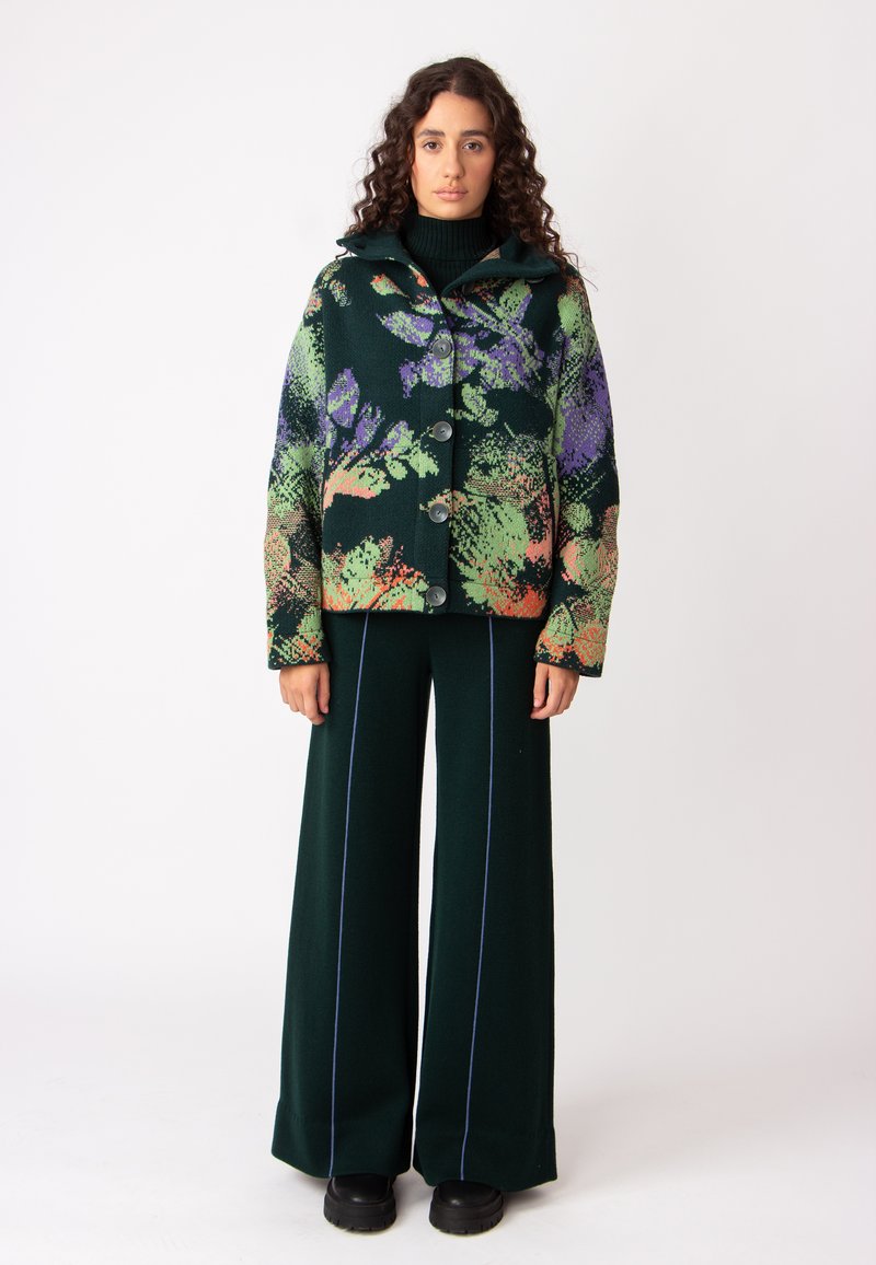 Jakna iz volnenega mešanega materiala v zeleni barvi s cvetličnim vzorcem v vijolični in oranžni barvi, z kapuco in velikimi gumbi, združena s širokimi zelenimi hlačami.
