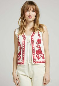 Gilet sans manches blanc avec des détails en broderie florale rouge et un bord festonné. Le gilet a un col en V et est fabriqué dans un tissu texturé.