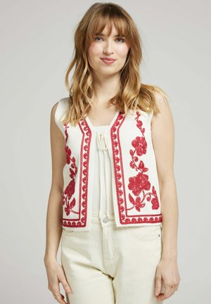 Gilet sans manches blanc avec des détails en broderie florale rouge et un bord festonné. Le gilet a un col en V et est fabriqué dans un tissu texturé.