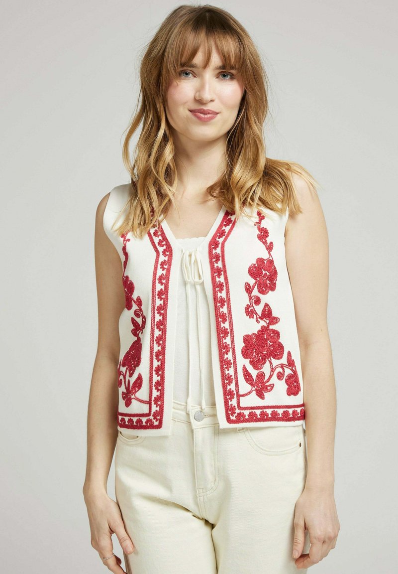 Gilet sans manches blanc avec des détails en broderie florale rouge et un bord festonné. Le gilet a un col en V et est fabriqué dans un tissu texturé.