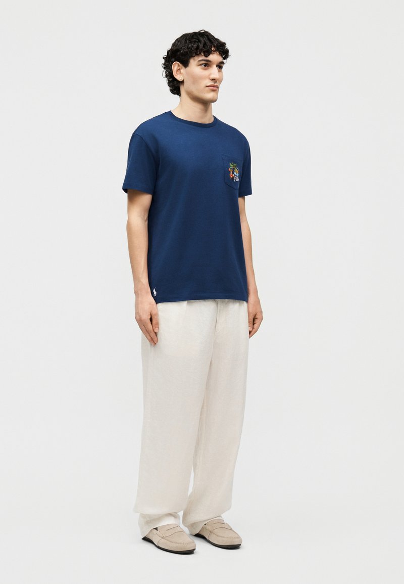 Man staat en draagt een marineblauw T-shirt met een grafisch zakontwerp, wijde witte broek en beige loafers, tegen een effen lichte achtergrond.