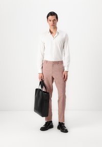 Un sac fourre-tout en cuir noir tenu par une personne portant une chemise blanche à boutons et un pantalon texturé pêche. Le tissu lisse et le design sur mesure sont visibles.