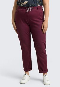 Pantalones color vino con cintura ajustable, puños enrollados y bolsillos laterales, combinados con zapatillas blancas. Textura de tela suave.