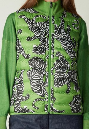 Grüne gesteppte Weste mit schwarz-weißem Tigerprint, getragen über einem grünen Langarmpullover und dunklen Jeans.