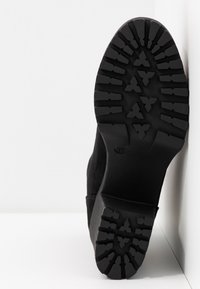 Semelle d'une botte noire avec une profondeur de crampons travaillés présentant des formes triangulaires et rectangulaires, positionnée verticalement contre un coin blanc.