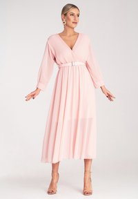 Jasnoróżowa długa sukienka z plisowanym topem, głębokim dekoltem w literę V, długimi rękawami i pasującym paskiem. Miękka, przezroczysta tkanina, długość do kostek z luźną spódnicą.