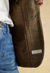 Tweed jakk, millel on pruun, roheline ja punane ruutpattern. Vooderdatud pruuni kangaga ja logotähisega, sisaldab see ühte nuppe.