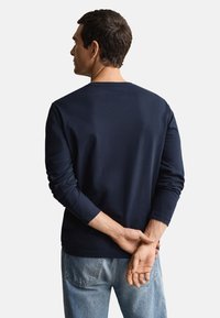 Marineblå langærmet T-shirt med rund hals, lavet af blødt bomuldsstof. Båret med lyseblå jeans, set bagfra.