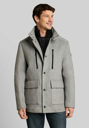 Mann trägt eine graue Jacke mit Reißverschluss, schwarzer Innenschicht und vorderen Klappentaschen, vor einem einfarbigen hellgrauen Hintergrund.