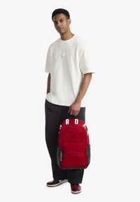 Ερυθρό σακίδιο πλάτης Jordan με μαύρες λεπτομέρειες, που διαθέτει έντονο λογότυπο, ρυθμιζόμενους ιμάντες και τσέπη με φερμουάρ στο μπροστινό μέρος. Φορεμένο με λευκό μπλουζάκι.