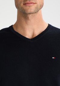 Uomo che indossa un maglione nero a V con un piccolo logo rosso, bianco e blu di Tommy Hilfiger sul petto.