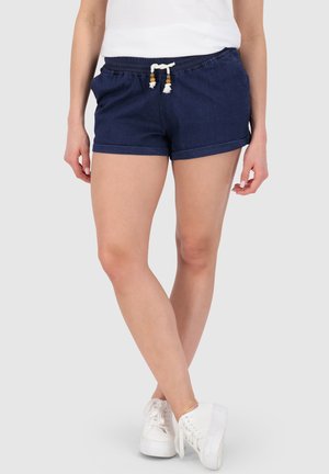 Frau trägt marineblaue Shorts mit Kordelzug, weiße Sneakers und ein weißes Oberteil, steht mit überkreuzten Beinen vor einem einfarbigen Hintergrund.