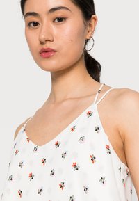 Camisola floral branca com alças finas, apresentando pequenos padrões de flores coloridas em vermelho, preto e azul, feita de um tecido leve.