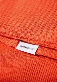 Pomarańczowa tkana tkanina o teksturowanej powierzchni, z białą etykietą z napisem "LINDERGH". Krawędzie są starannie zszyte.