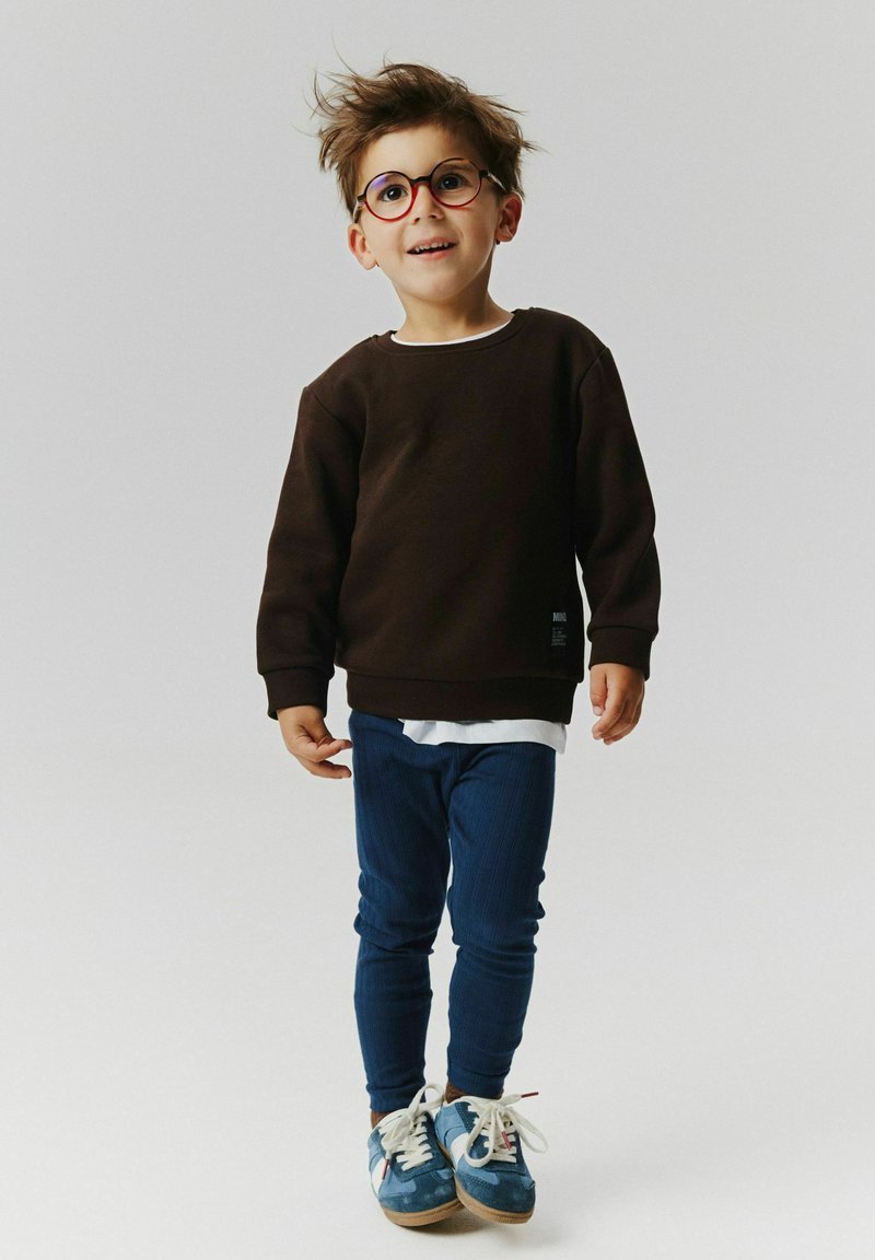 Jeune garçon avec des lunettes portant un sweat-shirt noir, un pantalon bleu et des baskets bleues, debout sur un fond blanc uni.