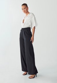 Witte blouse met korte mouwen, diepe V-hals en zak; zwarte hoge tailles brede broek; model draagt zwarte sandaal met bandjes.