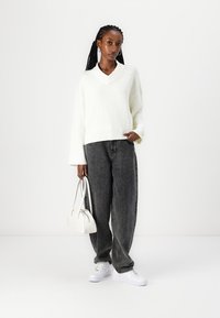 Hvit strikket genser med V-hals, kombinert med mørkegrå løse jeans. Modellen holder en liten hvit håndveske og har på seg hvite joggesko.