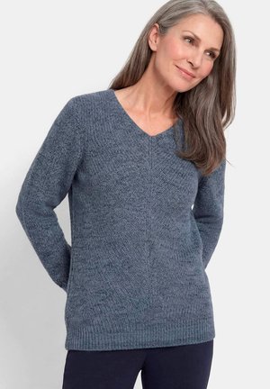 Femme aux longs cheveux gris portant un pull bleu en maille col en V et un pantalon foncé, debout les mains derrière le dos, souriant légèrement.