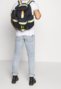 Zaino in tessuto nero con spallacci riflettenti gialli, molteplici tasche frontali e un logo distintivo. Indossato sulla schiena da una persona.